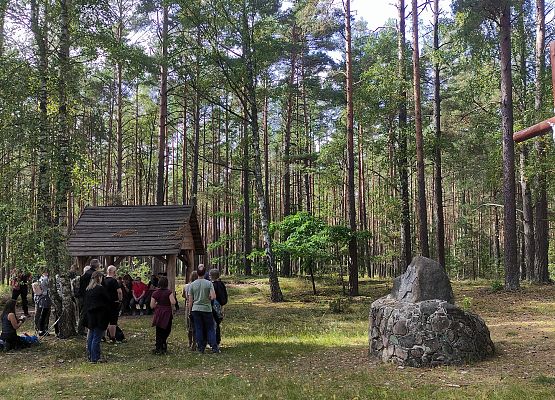 Przestrzeń, która łączy – spacer przez historię i krajobraz Grupa uczestników w lesie. Po prawej stronie pomnik Św. Huberta