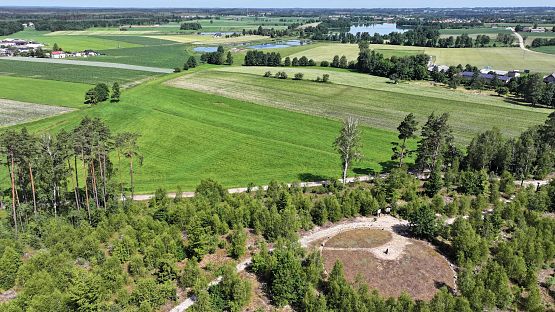 Panorama okolic Leśna. Na pierwszym planie krąg kamienny ze stelą fot. Mariusz Grzempa) Zdjęcie wykonane z góry na krąg kamienny, widok na zielone pola, w oddali jezioro i wieś Leśno.